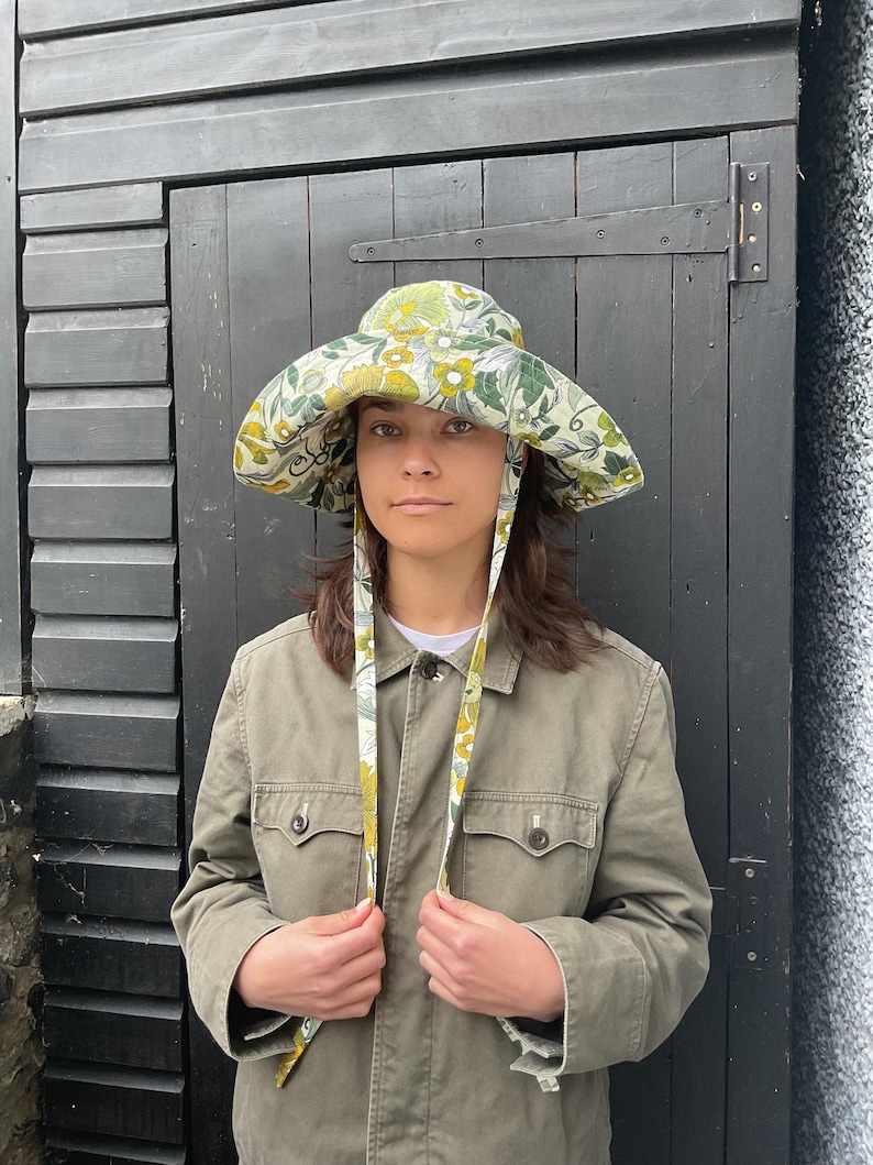 K&ouml;nnte beinhalten: Ein breitkrempiger Hut mit einem Blumenmuster in Gr&uuml;n-, Gelb- und Wei&szlig;t&ouml;nen. Der Hut hat lange B&auml;nder. Die Person im Bild tr&auml;gt eine olivgr&uuml;ne Jacke.