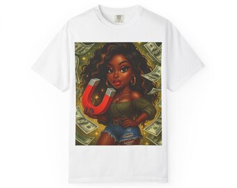 May include: White t-shirt featuring a colorful illustration of a woman holding a magnet with dollar bills in the background. The woman has dark skin, curly hair, and is wearing a green top and denim shorts. The magnet is red and silver.