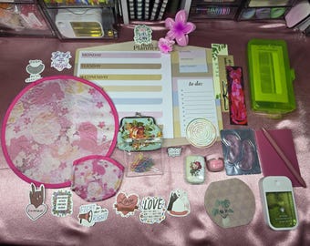 May include: A selection of stationery and accessories, including a weekly planner, stickers, a coin purse, and a pink fan. The items are arranged on a pink surface. Stickers have text like "Girls Can", "Love", and "Fight for your right".
