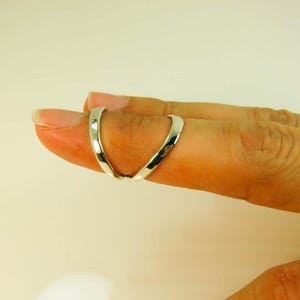 May include: A silver-colored ring designed to be worn on two fingers. The ring has a unique, curved design that wraps around the fingers. The ring is shown on a hand with a light skin tone, against a plain background.