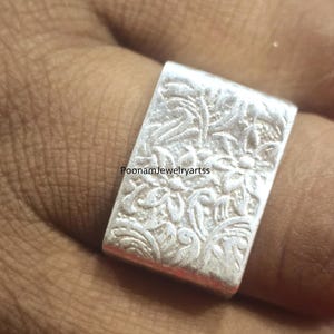 May include: A silver ring with a rectangular face featuring a floral pattern. The ring is a solid, bright silver color and appears to be handmade. The text "PoonamJewelryartss" is visible on the ring.