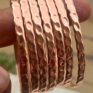 May include: A set of seven copper bangles with a hammered texture. The bangles are a warm, reddish-brown color and have a slightly irregular surface due to the hammering. The bangles are stacked together, showcasing their design.