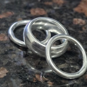 Könnte beinhalten: Ein Set aus drei silberfarbenen Ringen mit glatten, abgerundeten Oberflächen. Die Ringe variieren in der Dicke, wobei ein breites Band in der Mitte von zwei dünneren, kreisförmigen Bändern flankiert wird. Das Metall hat eine polierte Oberfläche.