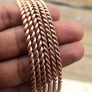 Puede incluir: Un conjunto de cinco pulseras de color cobre, de estilo trenzado. Las pulseras tienen un diseño de cuerda retorcida y se sostienen en una mano. Las pulseras son de un color marrón rojizo cálido.