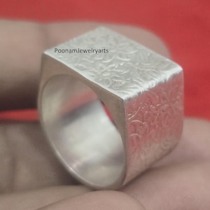 Op de afbeelding: Een zilveren ring met een rechthoekige bovenkant met een textuur bloemenontwerp. De ring wordt getoond tegen een rode achtergrond, met de tekst "PoonamJewelryarts" zichtbaar aan de bovenkant.