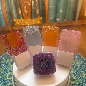 Op de afbeelding: Een verzameling decoratieve flessenstoppers in verschillende kleuren en ontwerpen. De stoppers zijn gerangschikt op een wit platform, met een paarse en iriserende stopper in het midden. Andere stoppers zijn rood, blauw, oranje en roze.