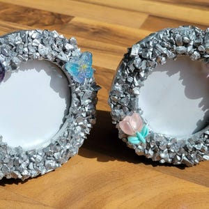 Op de afbeelding: Twee ovale fotolijsten met zilverkleurige, getextureerde randen. Elke lijst is versierd met decoratieve elementen, waaronder een paarse edelsteen, een blauwe vlinder en een roze bloem. De lijsten worden gepresenteerd op een houten oppervlak.