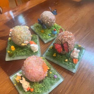 Puede incluir: Cuatro adornos decorativos de huevos de Pascua. Cada huevo está cubierto de purpurina y se asienta sobre un cuadrado de musgo verde. Conejitos, mariposas y pollitos en miniatura se colocan alrededor de los huevos. Los huevos son rosas, marrones y verdes.