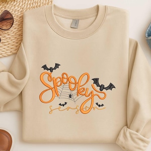 Könnte beinhalten: Beiges Sweatshirt mit dem Schriftzug "Spooky Season" in orangefarbener Schrift, mit Spinnennetz und Fledermäusen. Das Design ist auf der Vorderseite aufgestickt. Die Ärmel sind hochgekrempelt.