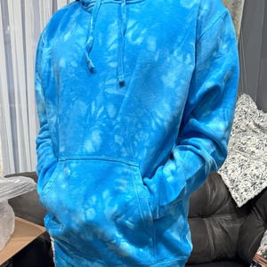 May include: A blue tie-dye hoodie with a front pocket and drawstring hood. The hoodie is a vibrant shade of blue with lighter blue tie-dye patterns throughout. The sleeves are long, and the garment appears to be made of a soft material.