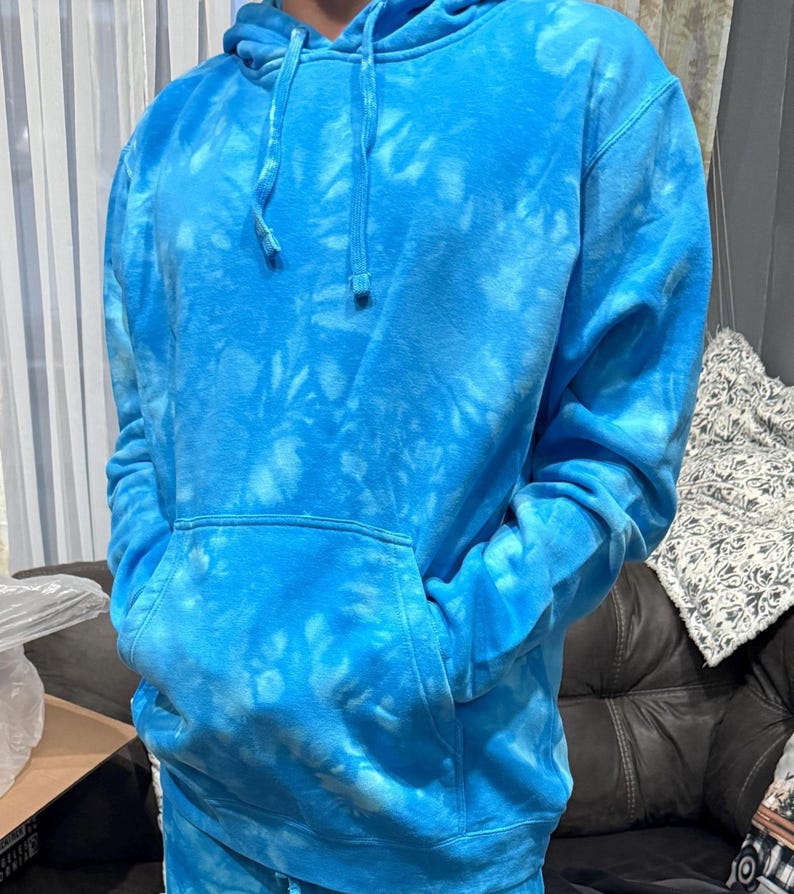 May include: A blue tie-dye hoodie with a front pocket and drawstring hood. The hoodie is a vibrant shade of blue with lighter blue tie-dye patterns throughout. The sleeves are long, and the garment appears to be made of a soft material.