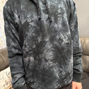 May include: A black and gray tie-dye hoodie with a drawstring hood and a kangaroo pocket. The sweatshirt has ribbed cuffs and hem. The tie-dye pattern creates a marbled effect across the fabric.