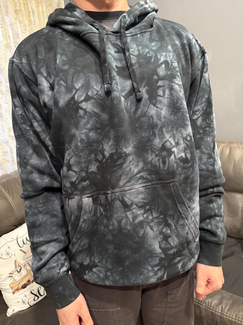 May include: A black and gray tie-dye hoodie with a drawstring hood and a kangaroo pocket. The sweatshirt has ribbed cuffs and hem. The tie-dye pattern creates a marbled effect across the fabric.