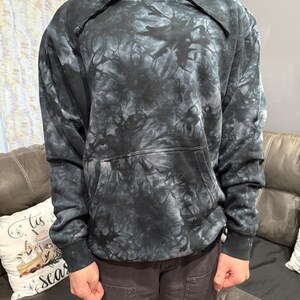 May include: A black and gray tie-dye hoodie with a drawstring hood and a front kangaroo pocket. The sweatshirt has long sleeves and ribbed cuffs and hem. The tie-dye pattern creates a marbled effect.