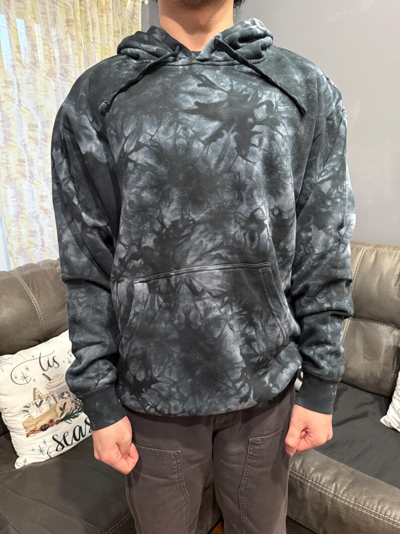 May include: A black and gray tie-dye hoodie with a drawstring hood and a front kangaroo pocket. The sweatshirt has long sleeves and ribbed cuffs and hem. The tie-dye pattern creates a marbled effect.