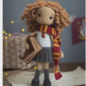 May include: Crocheted doll with curly brown hair, wearing a white shirt, gray skirt, and a red and gold scarf. The doll is holding a brown book and has gray socks and black shoes. The doll is a handmade item.