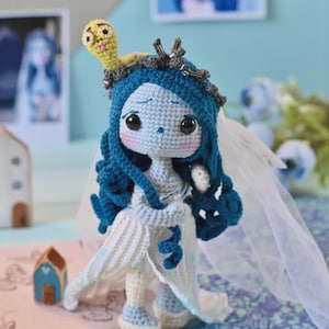 May include: A handmade crochet doll of a bride with blue hair and a white dress. The doll has large black eyes, rosy cheeks, and a small yellow worm on its head. The doll is wearing a veil and has a whimsical, cartoonish design.