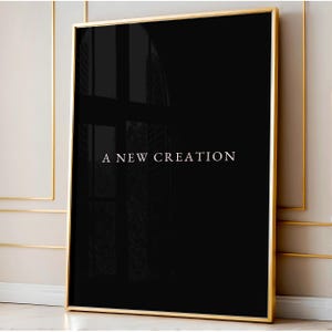 May include: A framed art print with a black background and the text "A NEW CREATION" in white. The frame is gold-coloured, and the artwork is displayed against a neutral-toned wall.