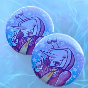 Puede incluir: Dos botones redondos con un personaje de dibujos animados con cabeza de televisor, nariz larga y micrófono. El personaje lleva un traje rojo con corbata amarilla. El fondo es azul claro con estrellas blancas.