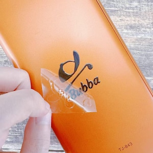 Può includere: Un oggetto rettangolare arancione con un logo nero "bba". Un adesivo trasparente viene staccato. Il testo "TJ-843" è stampato nell'angolo in basso a destra dell'oggetto.