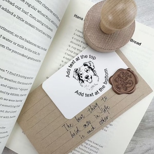 May include: A wooden stamp with a round top rests on a white card with a dog illustration and text. A brown wax seal and a kraft paper envelope are also visible, along with an open book.