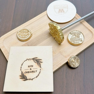 Puede incluir: Una colección de herramientas de marca personalizadas, que incluyen un hierro de marcar con mango de madera, varios sellos de latón y un posavasos de madera. El hierro de marcar tiene un diseño detallado. Un cuadrado de madera presenta el texto "RYE & CHEARLY" y un diseño floral.