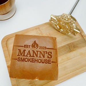 May include: A wooden cutting board with a rectangular piece of food branded with "EST 2023 MANN'S SMOKEHOUSE". A gold-colored branding iron is next to the food. A wooden cup with a dark liquid sits in the upper left corner.