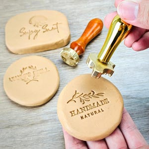 May include: A gold-colored wax seal stamp with a crown handle is pressed onto a tan wax circle, next to other wax seals and stamps. The stamped wax reads "HANDMADE NATURAL" with a leaf design. Other stamps and wax pieces are visible.