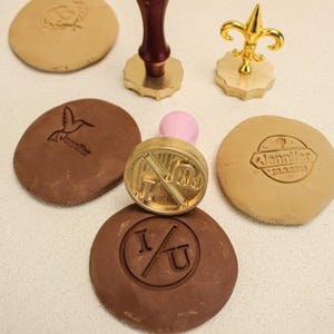 May include: An assortment of custom wax seal stamps and impressions. The stamps feature gold-colored metal heads with wooden handles and a fleur-de-lis design. Impressions include a hummingbird, the text "Jennifer", and the letters "I/U".