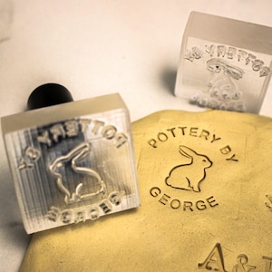 May include: Two clear, square pottery stamps featuring a bunny design and the text "Pottery by George." One stamp is pressed into clay, showing the embossed image. The stamps have a black handle.