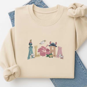 Pode incluir: Camisola bege com a palavra "ALOHA" bordada em cores pastel. O design inclui personagens de desenhos animados, uma flor de hibisco rosa e um sapo. A camisola é combinada com jeans azuis.