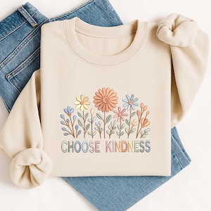 May include: A cream-coloured sweatshirt with a floral design featuring pastel-coloured flowers and the text "CHOOSE KINDNESS" in a rainbow font. The sweatshirt is folded on top of a pair of blue jeans.