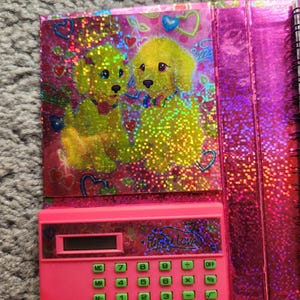 Puede incluir: Una calculadora rosa con botones verdes y el texto "Puppy Love". La calculadora forma parte de un cuaderno con una cubierta hologr&aacute;fica con dos cachorros amarillos y corazones.