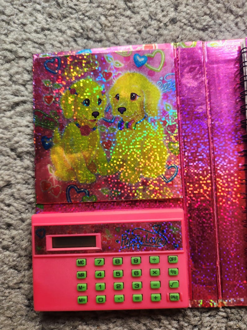 Puede incluir: Una calculadora rosa con botones verdes y el texto "Puppy Love". La calculadora forma parte de un cuaderno con una cubierta hologr&aacute;fica con dos cachorros amarillos y corazones.