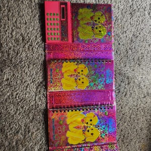 Puede incluir: Una calculadora rosa y tres cuadernos con espiral con un dise&ntilde;o hologr&aacute;fico y brillante. Los cuadernos y la calculadora presentan un gr&aacute;fico de cachorro amarillo repetido. Los cuadernos est&aacute;n etiquetados como "Diary-address book".
