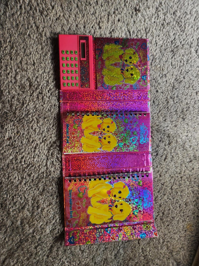 Puede incluir: Una calculadora rosa y tres cuadernos con espiral con un dise&ntilde;o hologr&aacute;fico y brillante. Los cuadernos y la calculadora presentan un gr&aacute;fico de cachorro amarillo repetido. Los cuadernos est&aacute;n etiquetados como "Diary-address book".