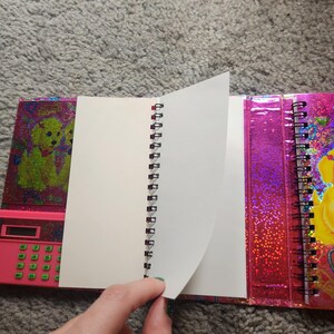 Puede incluir: Un cuaderno con espiral con una cubierta rosa que presenta una calculadora y un gr&aacute;fico de perro. Las p&aacute;ginas del cuaderno son blancas y la cubierta tiene un acabado hologr&aacute;fico y brillante. El cuaderno est&aacute; abierto, con una mano pasando una p&aacute;gina.
