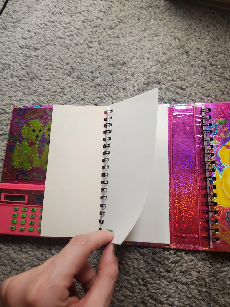 Puede incluir: Un cuaderno con espiral con una cubierta rosa que presenta una calculadora y un gr&aacute;fico de perro. Las p&aacute;ginas del cuaderno son blancas y la cubierta tiene un acabado hologr&aacute;fico y brillante. El cuaderno est&aacute; abierto, con una mano pasando una p&aacute;gina.