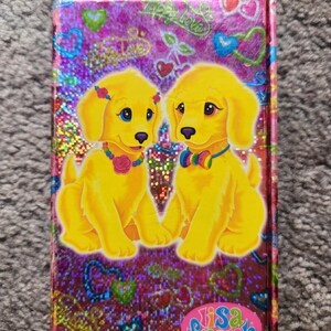Puede incluir: Un cuaderno de Lisa Frank con una cubierta hologr&aacute;fica. Presenta dos cachorros de dibujos animados amarillos con ojos azules, con collares coloridos y flores. El fondo es brillante con corazones y las palabras "Happy Love". El logotipo de Lisa Frank est&aacute; en la parte inferior.