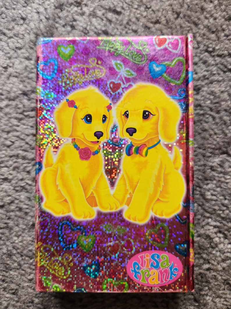 Puede incluir: Un cuaderno de Lisa Frank con una cubierta hologr&aacute;fica. Presenta dos cachorros de dibujos animados amarillos con ojos azules, con collares coloridos y flores. El fondo es brillante con corazones y las palabras "Happy Love". El logotipo de Lisa Frank est&aacute; en la parte inferior.