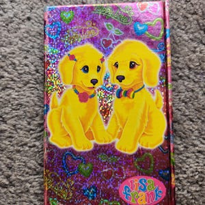 Puede incluir: Un cuaderno de Lisa Frank con una cubierta morada brillante, con dos cachorros amarillos de dibujos animados con ojos azules, que llevan collares coloridos y accesorios florales. La portada tiene corazones y la frase "Happy Love".