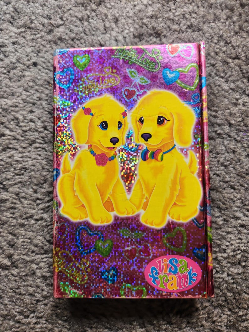 Puede incluir: Un cuaderno de Lisa Frank con una cubierta morada brillante, con dos cachorros amarillos de dibujos animados con ojos azules, que llevan collares coloridos y accesorios florales. La portada tiene corazones y la frase "Happy Love".