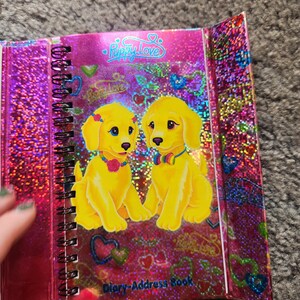 Puede incluir: Un diario-libro de direcciones con espiral y cubierta rosa brillante. La portada presenta dos cachorros de dibujos animados amarillos con ojos azules, con collares y lazos. El texto "Puppy Love" est&aacute; en la parte superior y "Diary-Address Book" en la inferior.