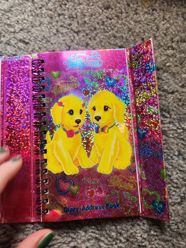 Puede incluir: Un diario-libro de direcciones con espiral y cubierta rosa brillante. La portada presenta dos cachorros de dibujos animados amarillos con ojos azules, con collares y lazos. El texto "Puppy Love" est&aacute; en la parte superior y "Diary-Address Book" en la inferior.