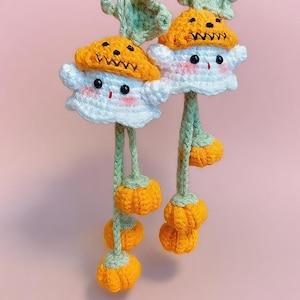 May include: Two handmade crochet mushroom ornaments. Each ornament features a white ghost-like body with a smiling face, an orange cap, and dangling orange pumpkin-shaped accents. The stems and leaves are green. The background is a solid pink.