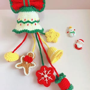 May include: A handmade Christmas bell ornament with a red bow and green trim. The bell is white with red and green accents. Dangling from the bell are crocheted yellow stars, a gingerbread man, a red flower, and a yellow bell. A snowman and Santa Claus ornament are also present.