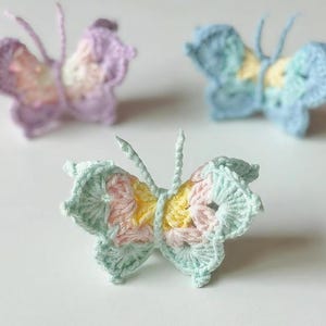 May include: Three handmade crochet butterflies in pastel colors. The butterflies have intricate wing patterns in shades of pink, yellow, blue, and purple. The butterflies are displayed on a white surface.