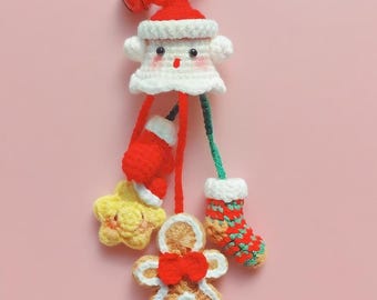 May include: A handmade Christmas-themed keychain featuring a crocheted Santa Claus figure with a red hat and a bell. The keychain includes a red stocking, a yellow star, and a gingerbread man, all in festive colors. The background is a solid pink.