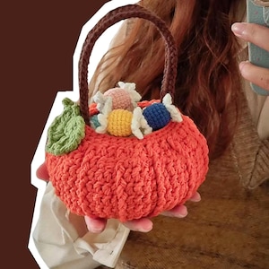 May include: An orange crochet pumpkin basket with a brown handle, filled with colourful crochet sweets. A green leaf is attached to the side. The basket is held in a person's hands.