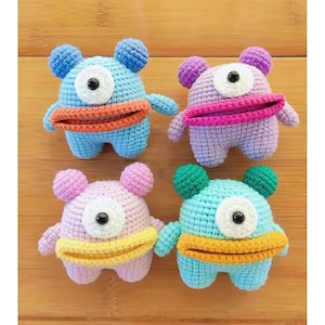 May include: Four colorful crochet monster toys. Each has a single eye, small arms, and a wide mouth. The monsters are in shades of blue, purple, pink, and green, with contrasting mouth and ear colors. These handmade toys are perfect for children.